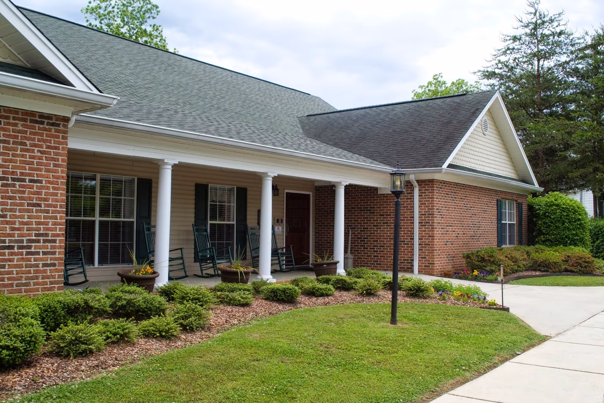 Front exterior of a single-story brick senior living building with a covered porch, white columns, rocking chairs and landscaped lawn.