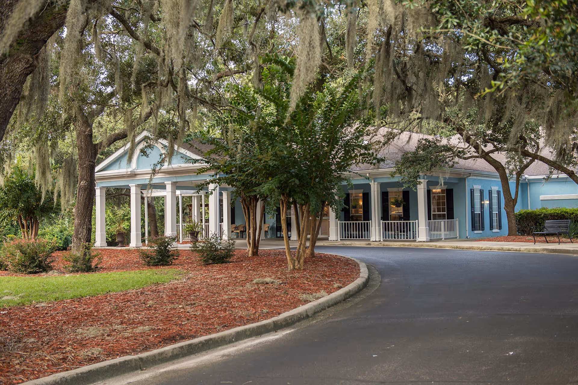 Front exterior of a blue single-story senior living building with a columned porch, trees with hanging Spanish moss, and a curved driveway.