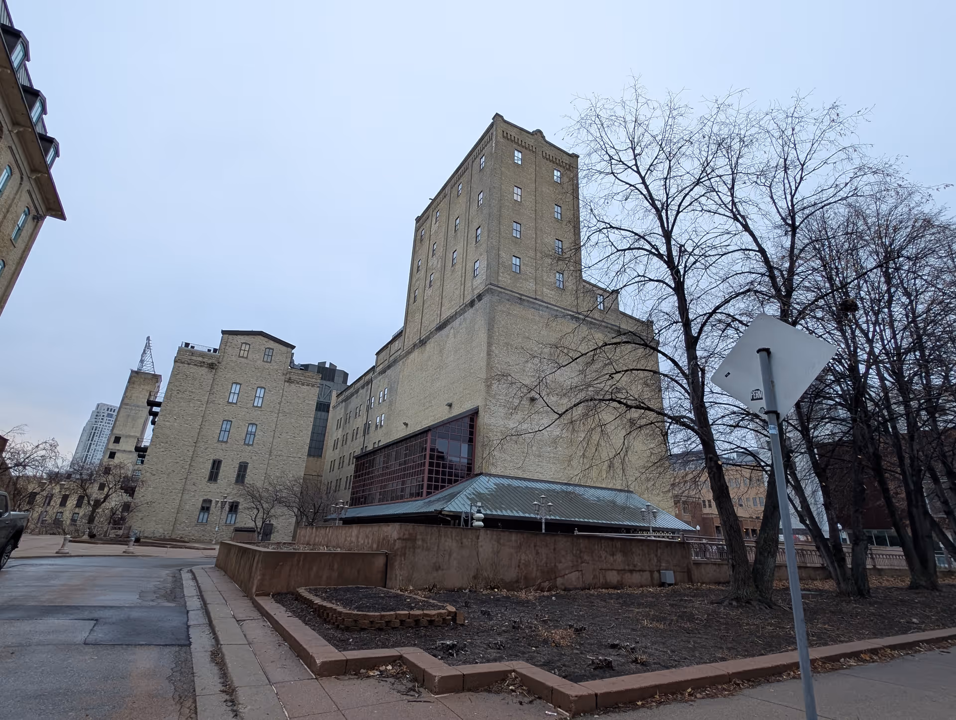 Large beige brick multi-story building viewed from the street with leafless trees and a sidewalk under an overcast sky.
