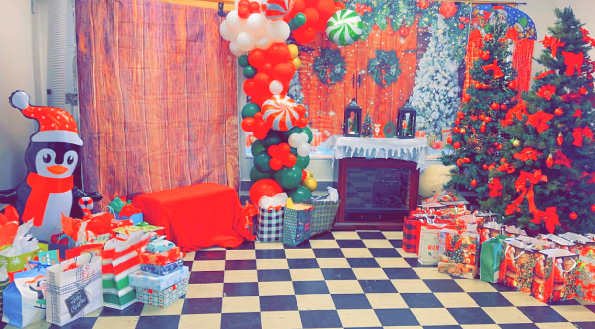Festively decorated indoor scene with Christmas trees, wrapped gifts, a balloon arch, a penguin prop and a faux fireplace on a black-and-white tiled floor.