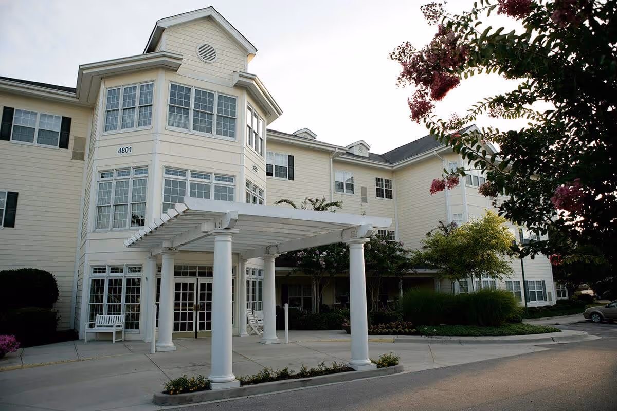 Front entrance of a light-colored multi-story senior living building with large windows, white columns and a covered porte-cochere.