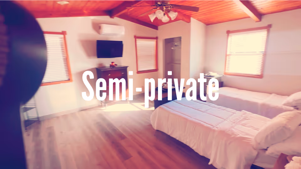 A semi-private bedroom with two beds, wooden ceiling, blinds, dresser, and a wall-mounted TV.
