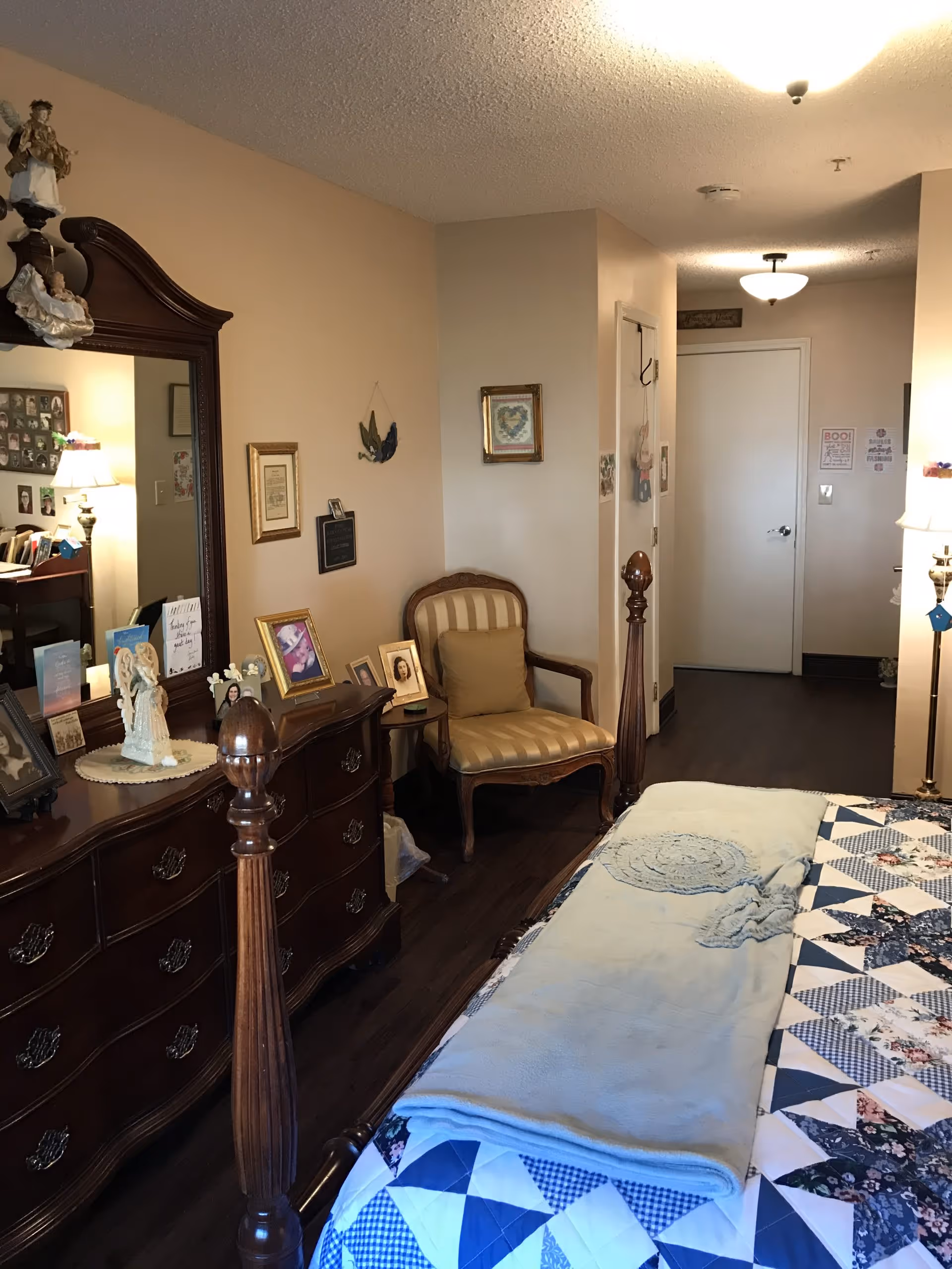A cozy bedroom in a senior living community featuring a wooden bed with a quilted bedspread and a light blue blanket. There is a wooden dresser with a large mirror on the left side, adorned with framed photos and decorative items. A cushioned armchair with a pillow is placed against the wall near the door. The room has warm beige walls and dark wood flooring, with soft lighting from a floor lamp on the right.