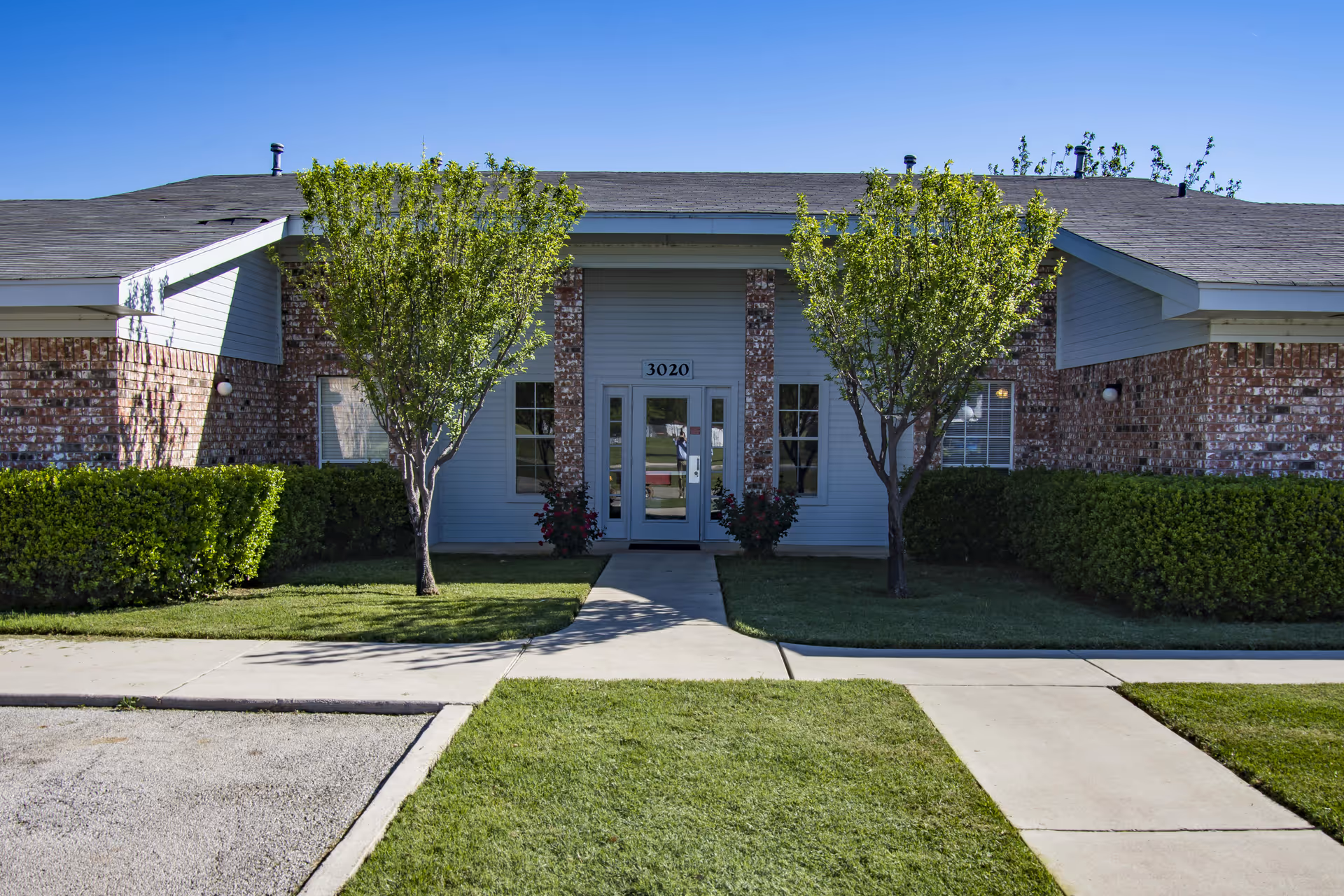 Front entrance of a low brick building with double glass doors numbered 3020, flanked by two small trees and trimmed hedges.