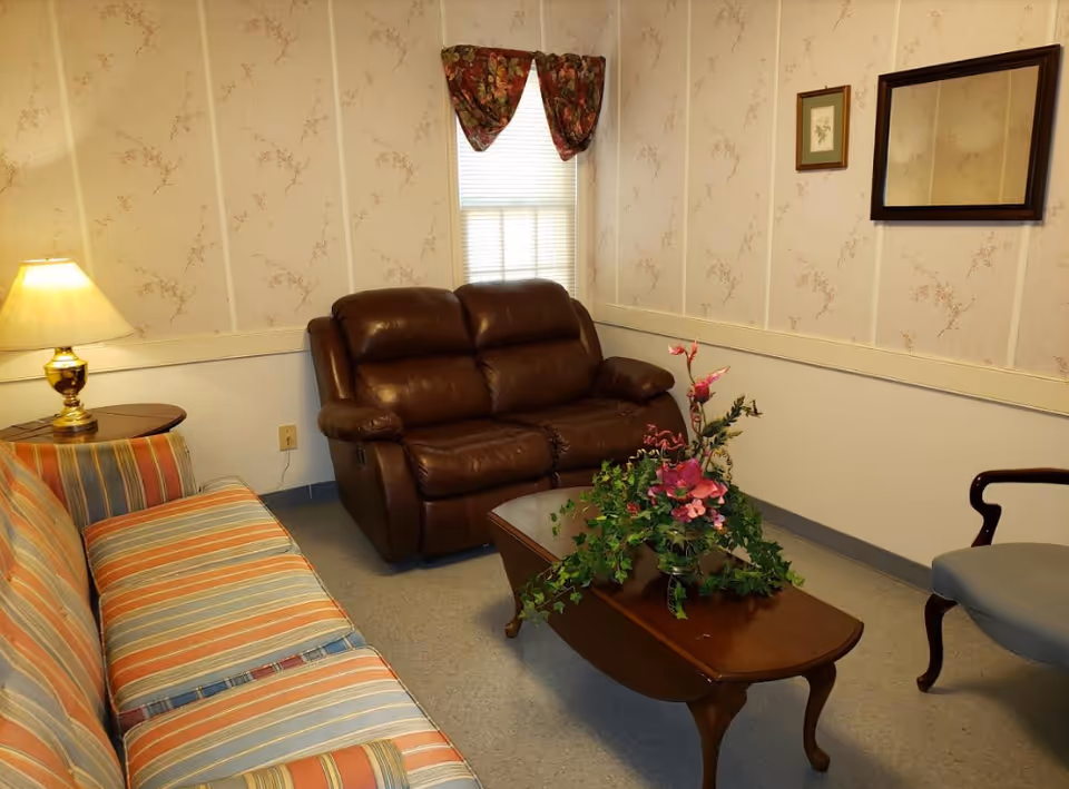 A cozy living room with striped sofa, leather recliner, coffee table, flowers, and a lamp.