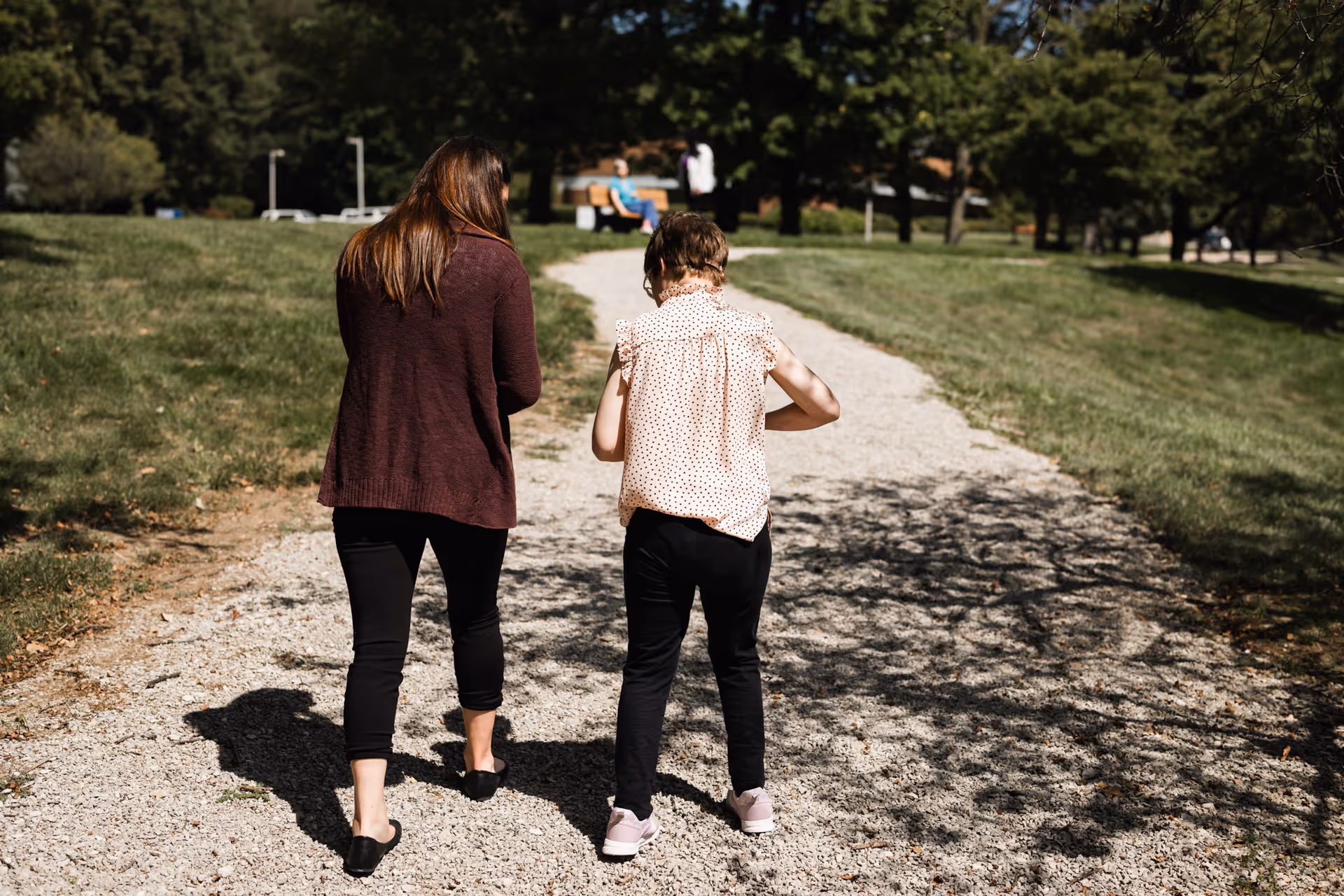 Two people walk along a gravel path in a sunny park with benches.