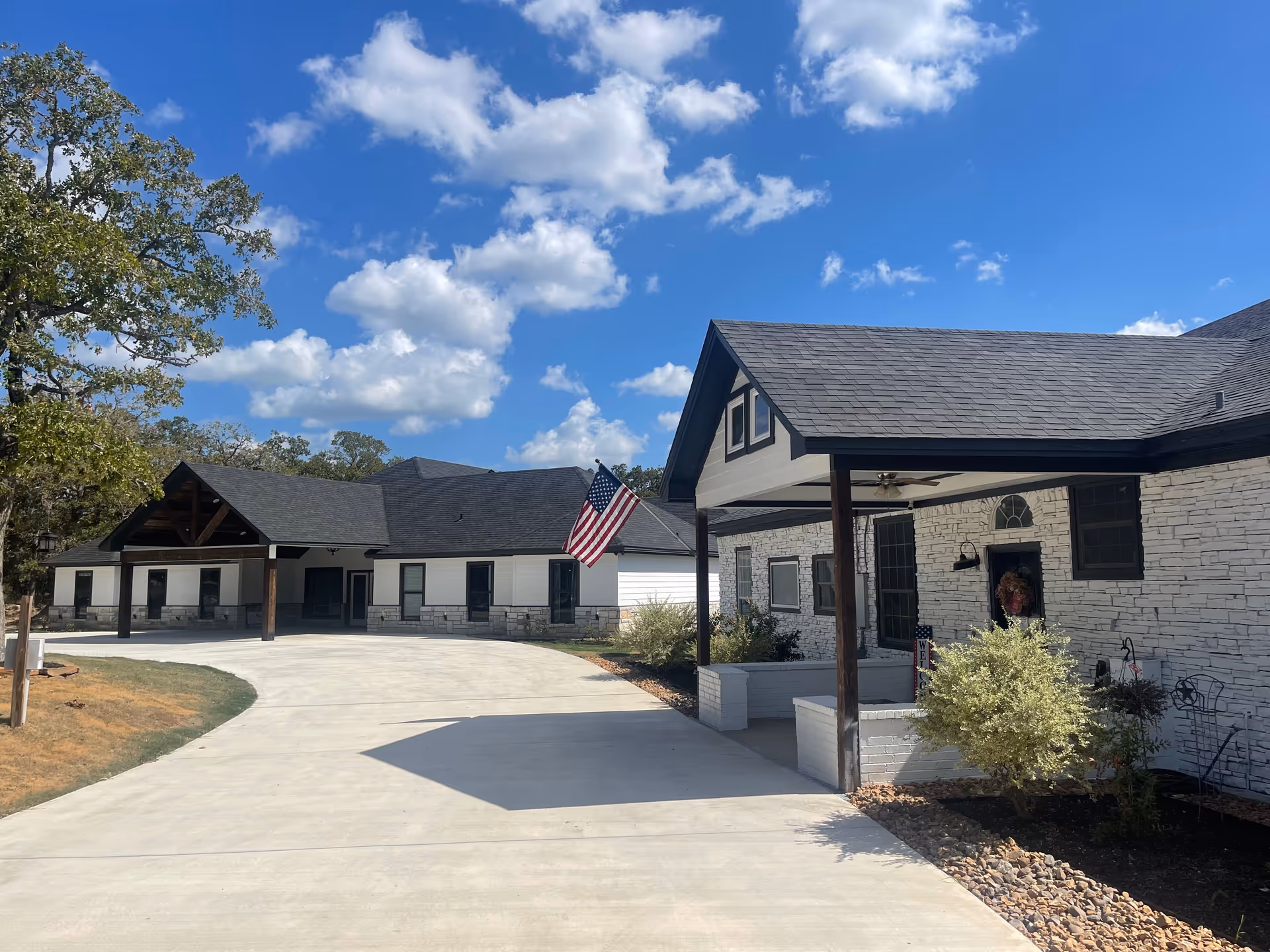 Exterior view of a single-story assisted living facility building with white stone walls and dark shingled roofs under a blue sky with scattered clouds. An American flag is mounted near the entrance, and there is a concrete driveway leading up to the building. Some shrubs and trees are visible around the building.