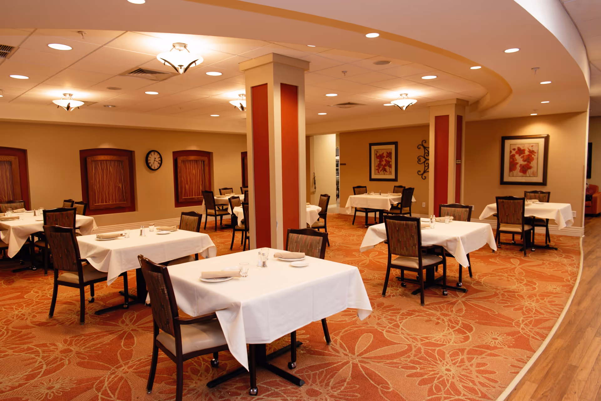 A warm dining room with multiple tables covered in white tablecloths and chairs arranged around columns and framed artwork on the walls.