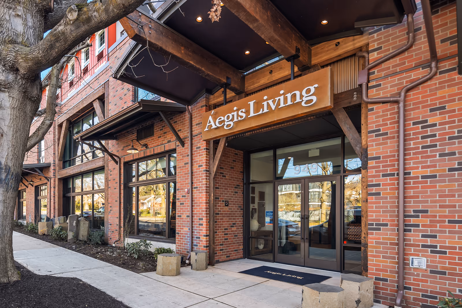 Brick facade and main entrance of the Aegis Living building with a wooden awning, glass doors, and a sidewalk.