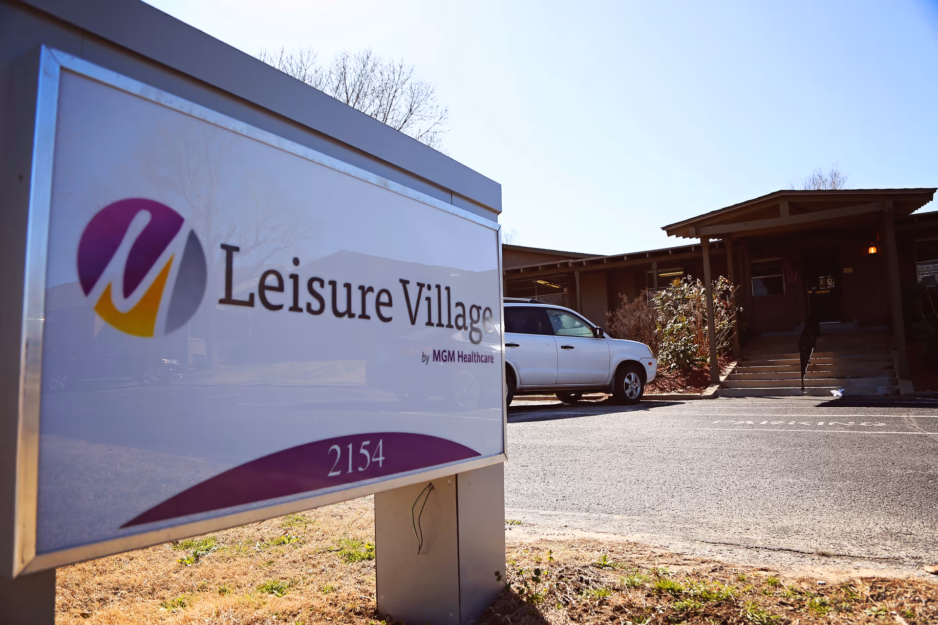 Leisure Village sign in foreground with a parked white car and building entrance behind.