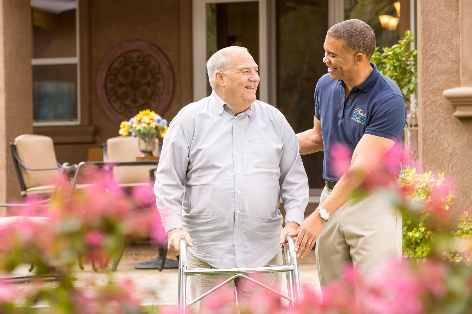 Caregiver assists an elderly man with a walker in a sunny outdoor courtyard.
