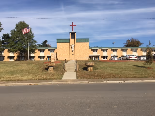 Front view of a two-story yellow brick care facility with a central tower topped by a red cross, a walkway and lawn, and an American flag.