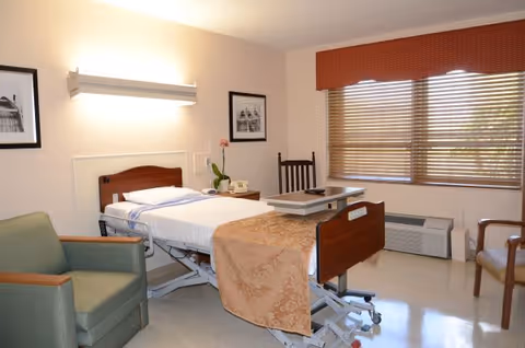 A hospital-style patient bedroom with a bed, desk, chair, and window blinds.