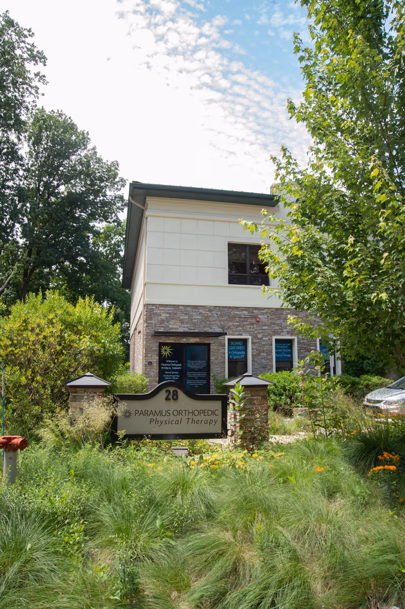Two-story building entrance with stone facade, sign Paramus Orthopedic Physical Therapy, surrounded by greenery.