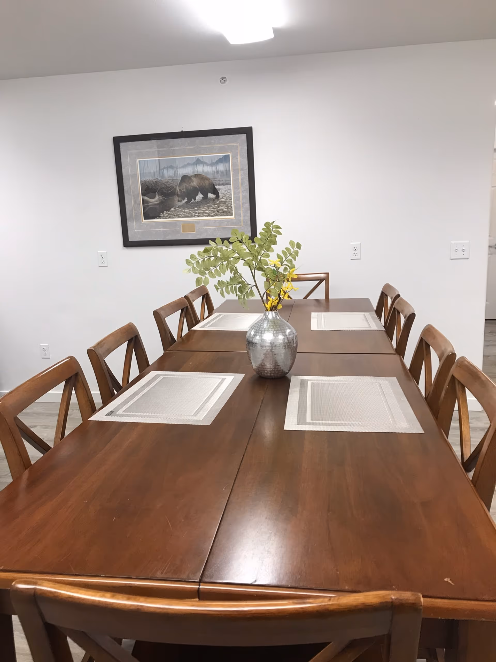 A long wooden dining table with placemats, chairs, and a vase in a bright dining room.