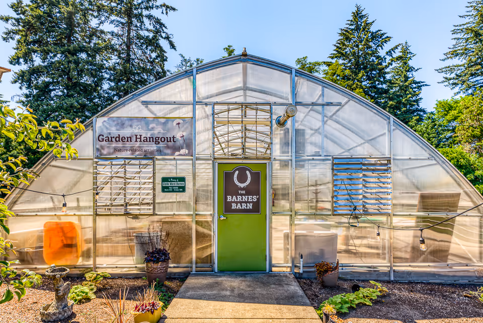 Front entrance of a greenhouse-style space labeled 'The Barnes' Barn' and 'Garden Hangout' with a green door and surrounding plants.