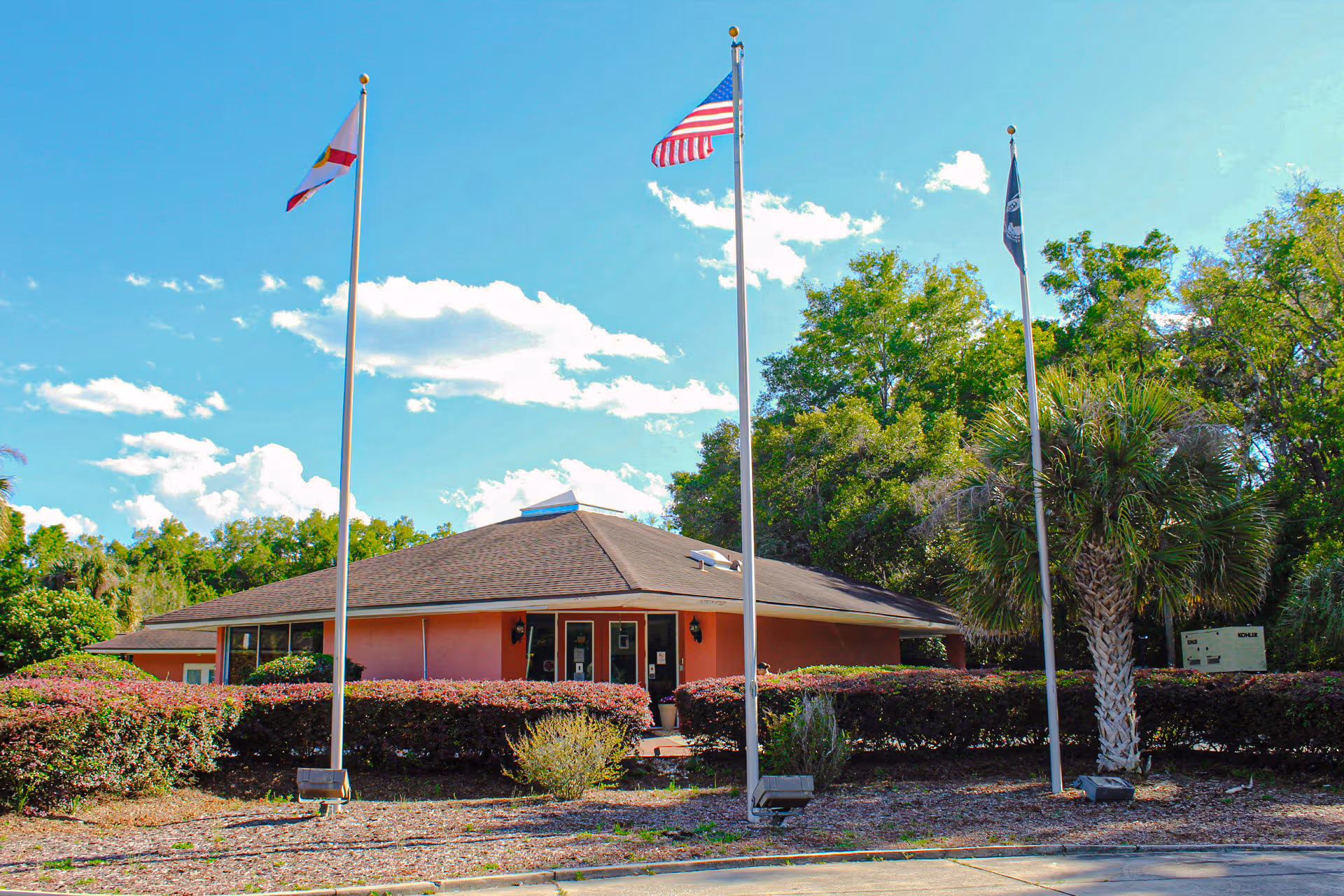 A pinkish-orange, single-story building with three flagpoles and trimmed hedges in front.