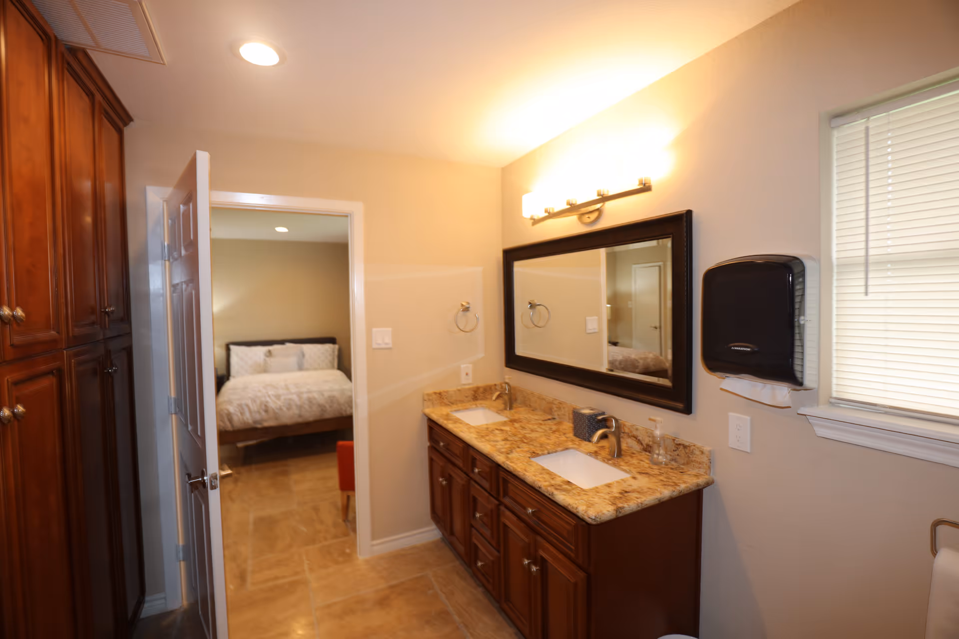 Bright bathroom with double vanity, granite countertop, mirror, and doorway to a bedroom.