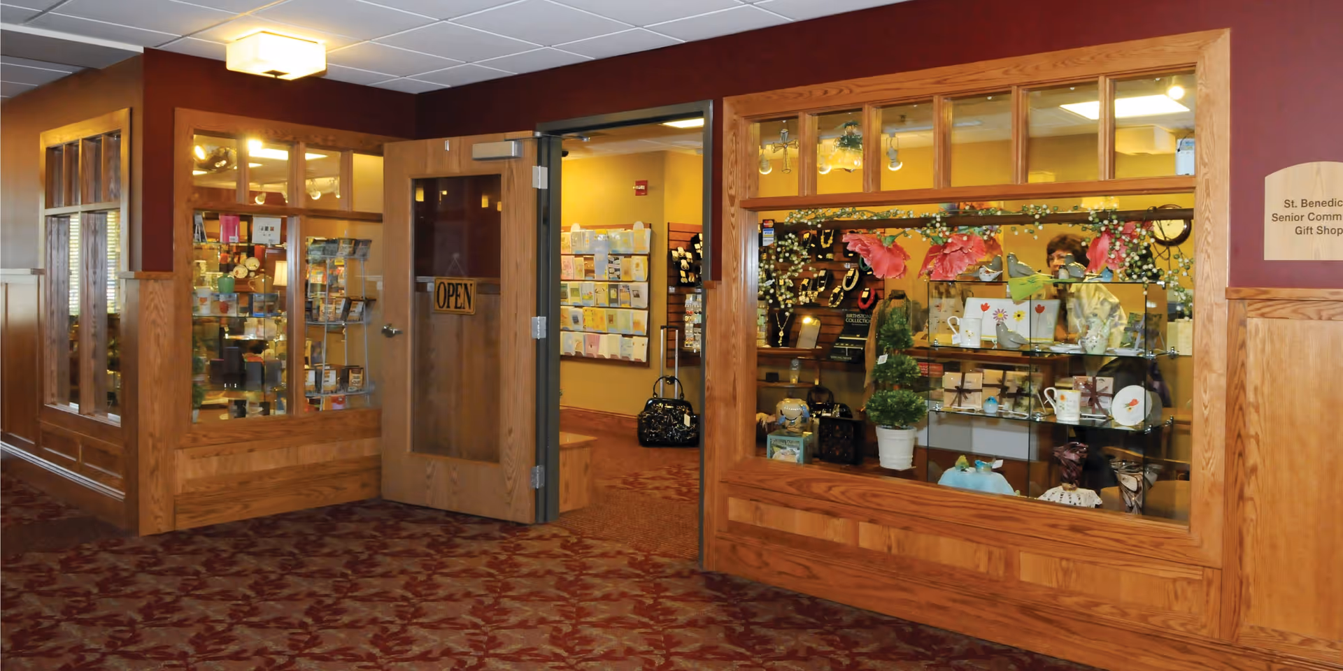 Interior view of a senior community gift shop with wooden framed glass windows and door. The door is open with an 'OPEN' sign, revealing shelves with greeting cards and various gift items. The walls are painted in warm tones with a patterned carpet on the floor.