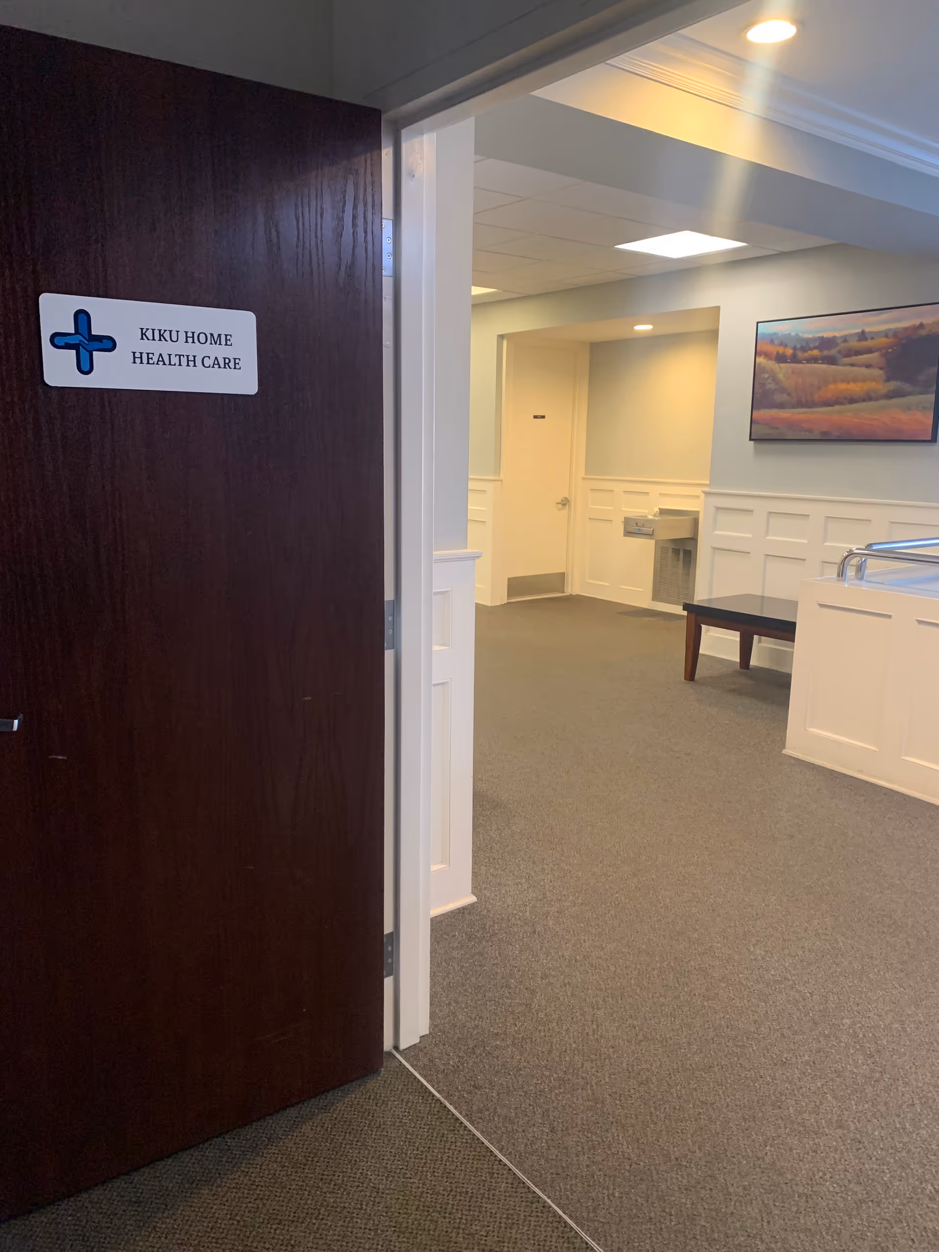 Interior corridor of a facility with doorway, wainscoted walls, and a KIKU HOME HEALTH CARE sign.
