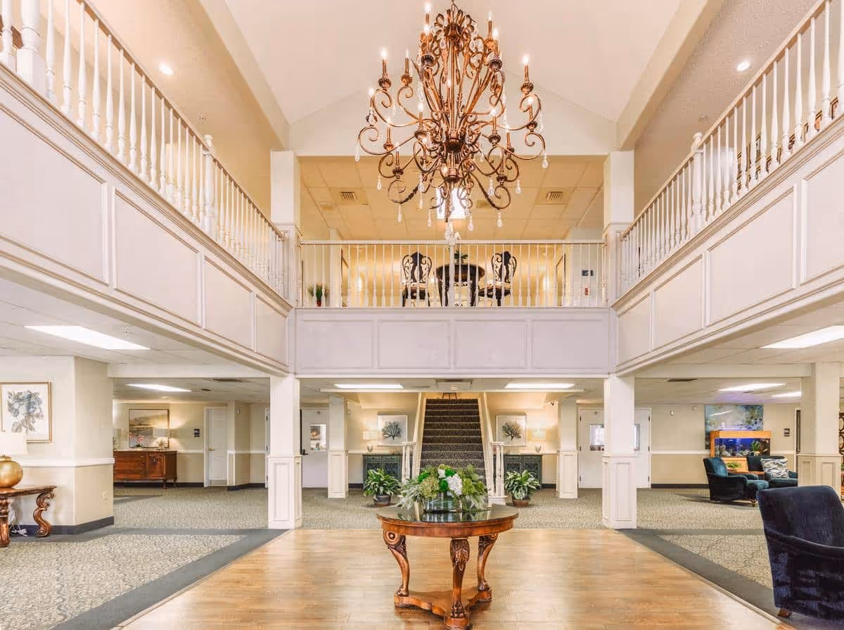 Spacious and elegant senior living facility lobby with a large ornate chandelier hanging from a high ceiling, a round wooden table with a floral arrangement in the center, carpeted floors with seating areas on the sides, and a staircase leading to an upper balcony with white railings.