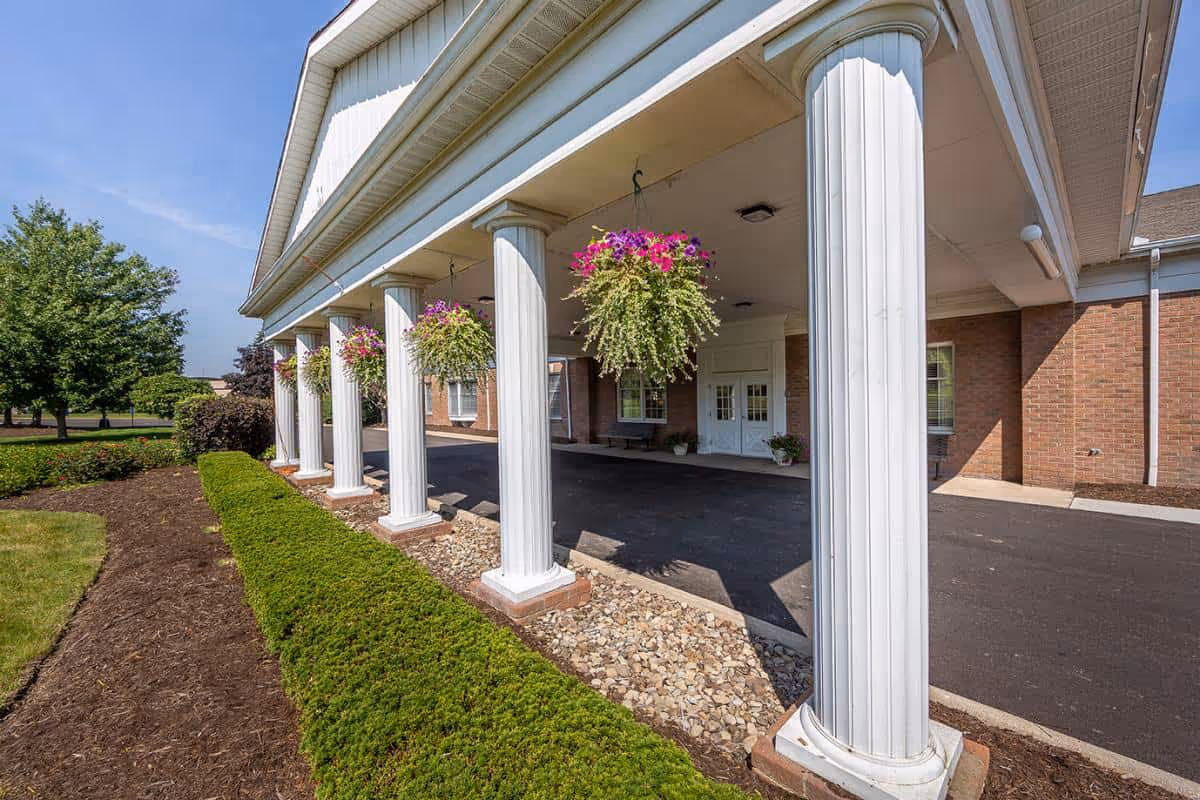 Entrance of a brick building with white columns supporting a covered driveway. Hanging flower baskets with pink and white flowers are suspended from the ceiling. There is a neatly trimmed hedge and landscaped garden bed along the side of the driveway. The sky is clear and blue.