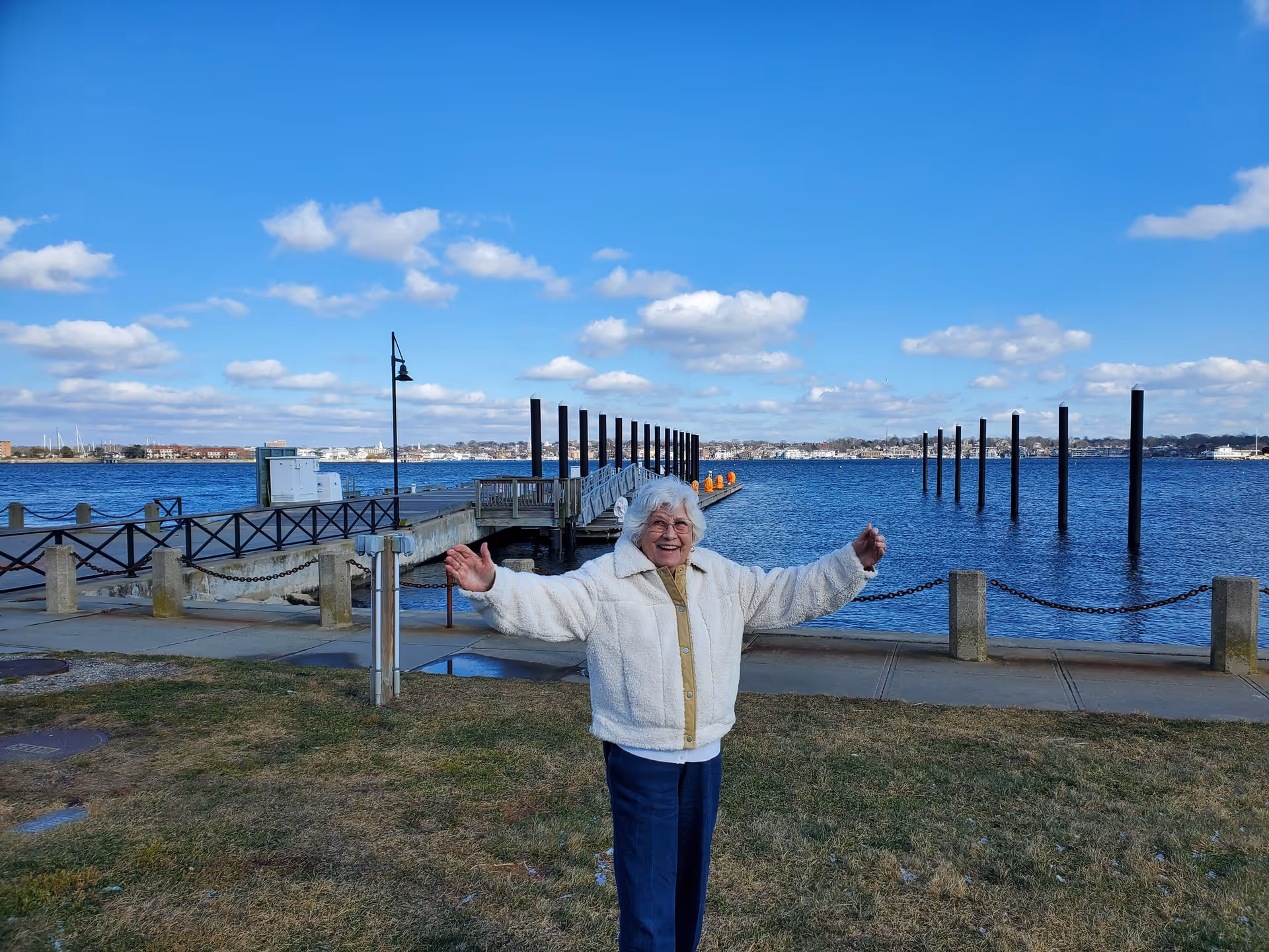 An elderly woman wearing a white jacket and dark pants stands on a grassy area near a waterfront with her arms outstretched. Behind her is a pier extending into the water, with a clear blue sky and scattered clouds above.