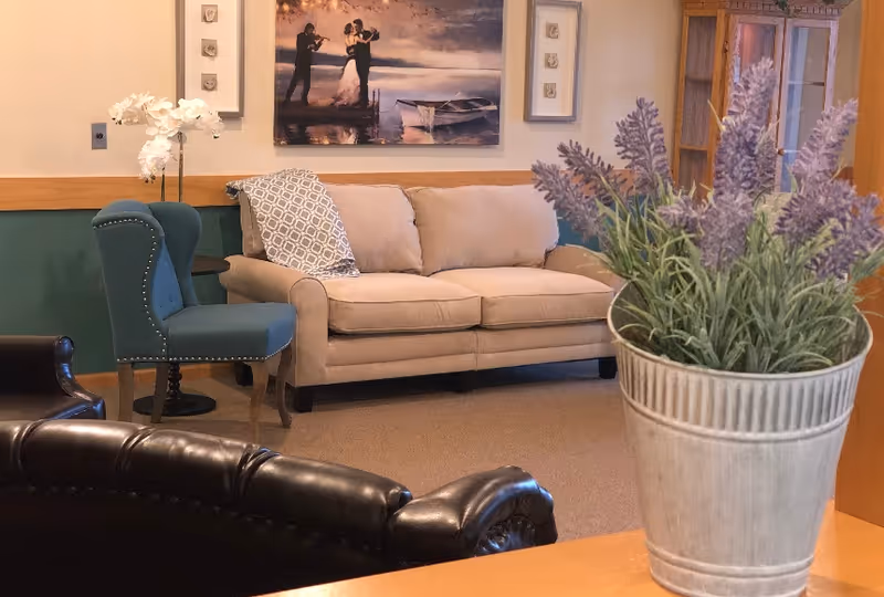 A cozy living room area with a beige sofa, a blue upholstered chair, and a dark brown leather chair. A framed painting of a couple dancing near a boat hangs on the wall above the sofa. A potted plant with purple flowers is in the foreground on a wooden surface. The room has warm lighting and a calm, inviting atmosphere.