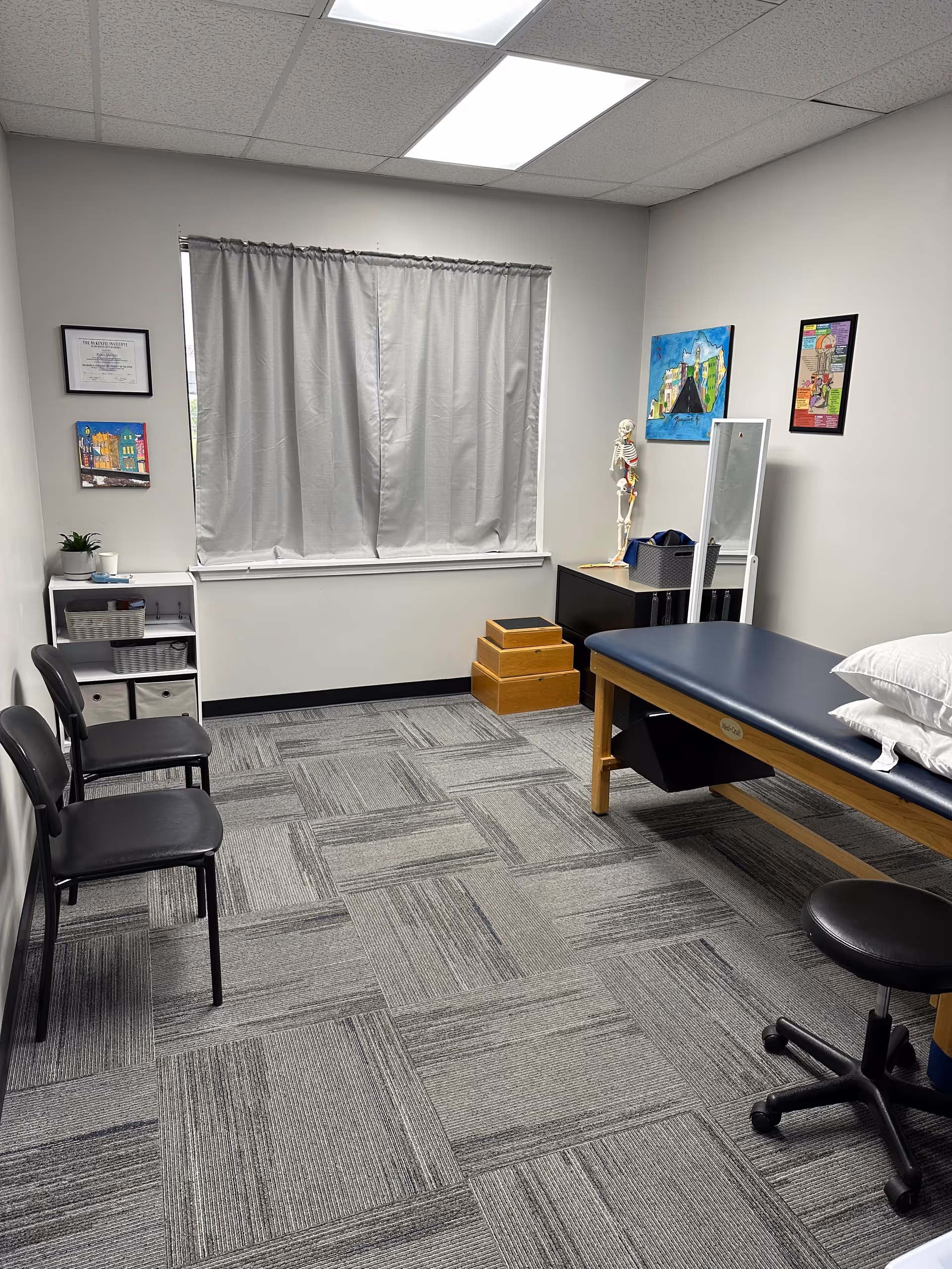 Physical therapy examination room with a table, skeleton model, chairs, and window curtains.