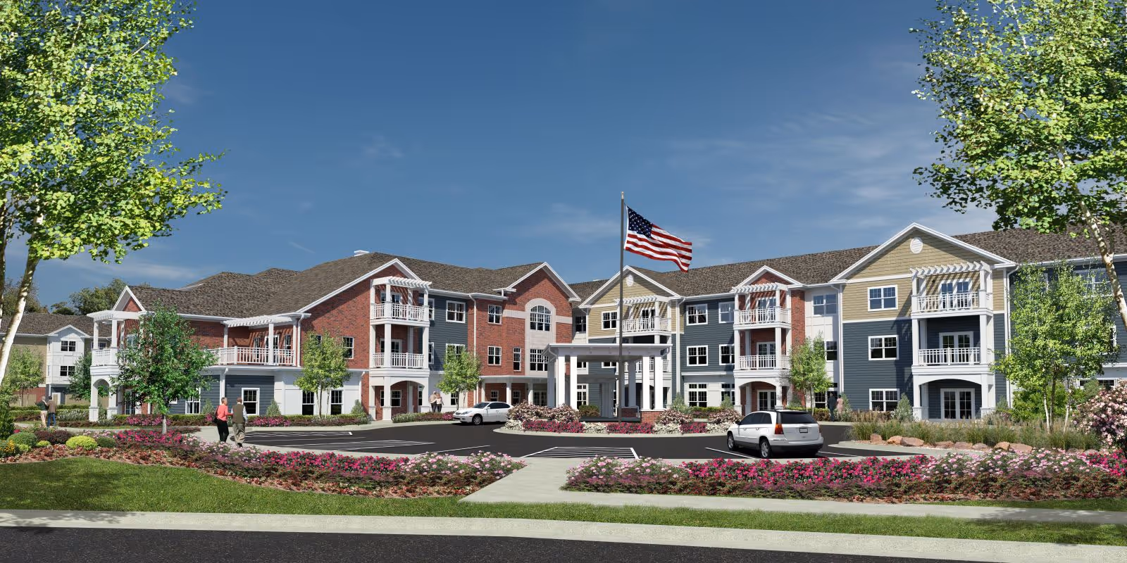 Exterior view of a multi-story senior living facility with a mix of brick and siding facades, balconies, and a central entrance with a covered drop-off area. The building is surrounded by landscaped gardens with flowers, trees, and a flagpole flying the American flag. A few cars are parked in front and a person is walking on the sidewalk.