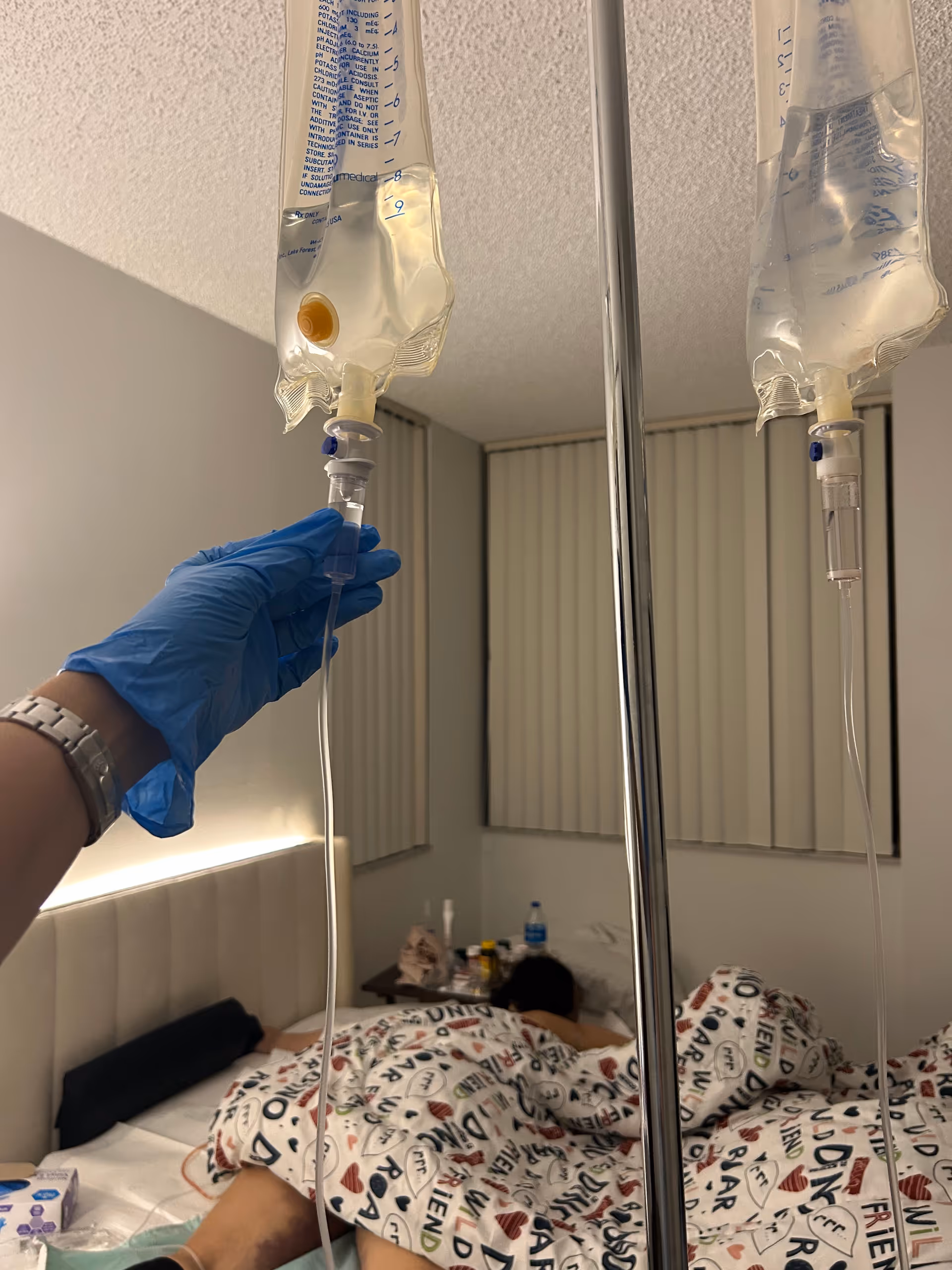 Gloved caregiver adjusts IV bag lines beside a patient in bed.