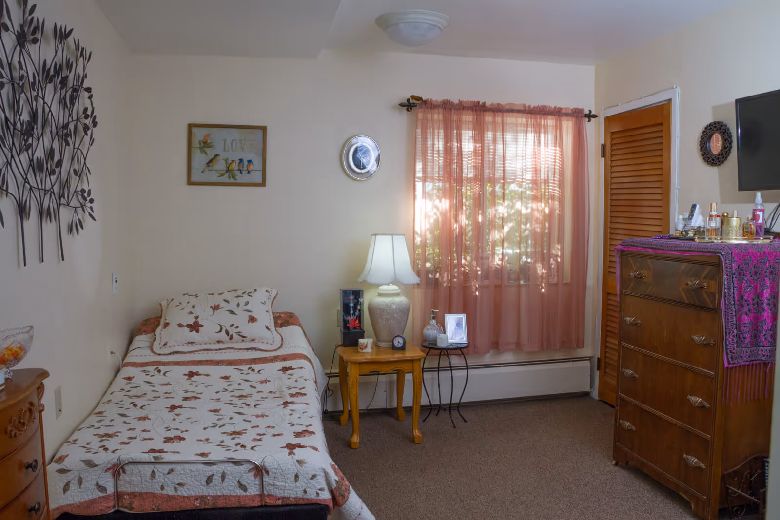 A cozy bedroom with a single bed covered in a floral quilt. Next to the bed is a wooden nightstand with a lamp, clock, and picture frame. A window with sheer pink curtains lets in natural light. On the right side, there is a wooden dresser with a purple cloth on top and various personal items. The walls are decorated with a metal tree wall art and framed pictures.