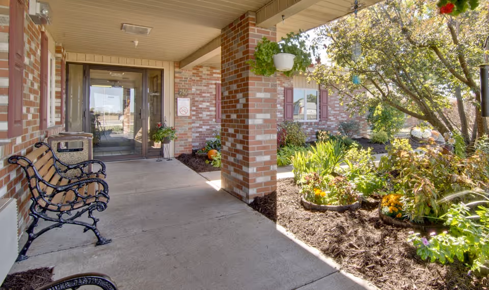 Covered brick entrance walkway with benches, a glass door entry, and landscaped flowerbeds.