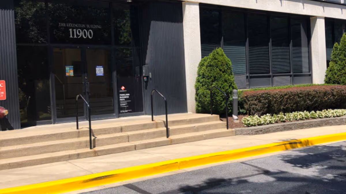 Exterior view of a building entrance with steps, glass doors, and landscaped shrubs.