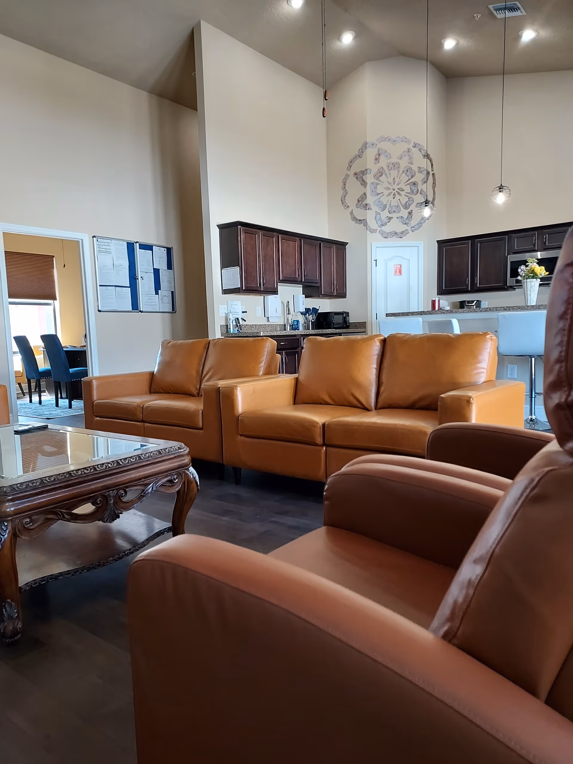Cozy senior living lounge with tan leather seating, a glass coffee table, and an open kitchen.