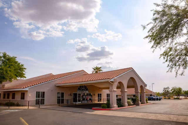 Exterior view of a single-story building with a tiled roof and arched entrance, surrounded by a parking lot and some trees under a partly cloudy sky.