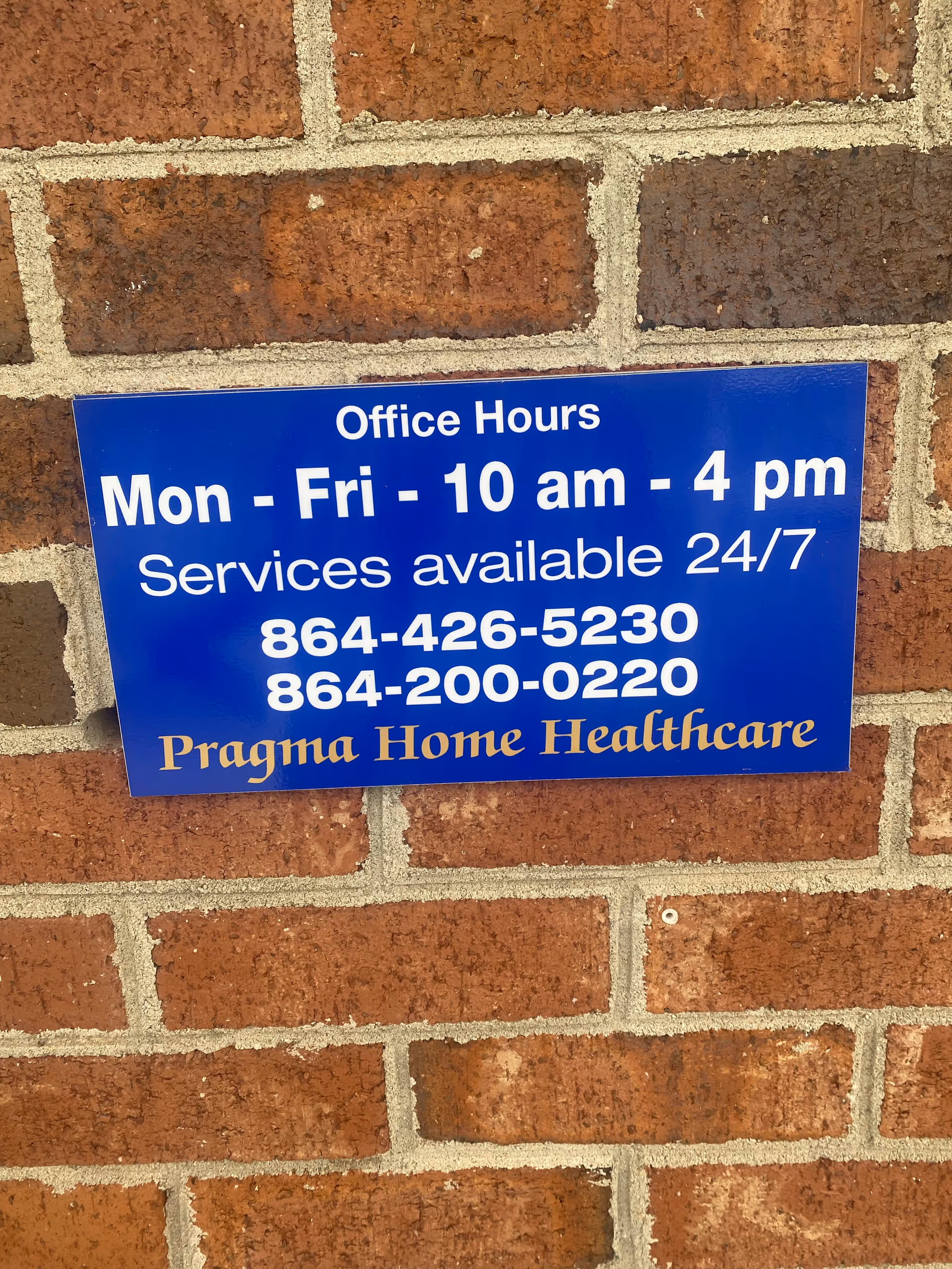 Blue sign on brick wall showing office hours and Pragma Home Healthcare contact numbers.