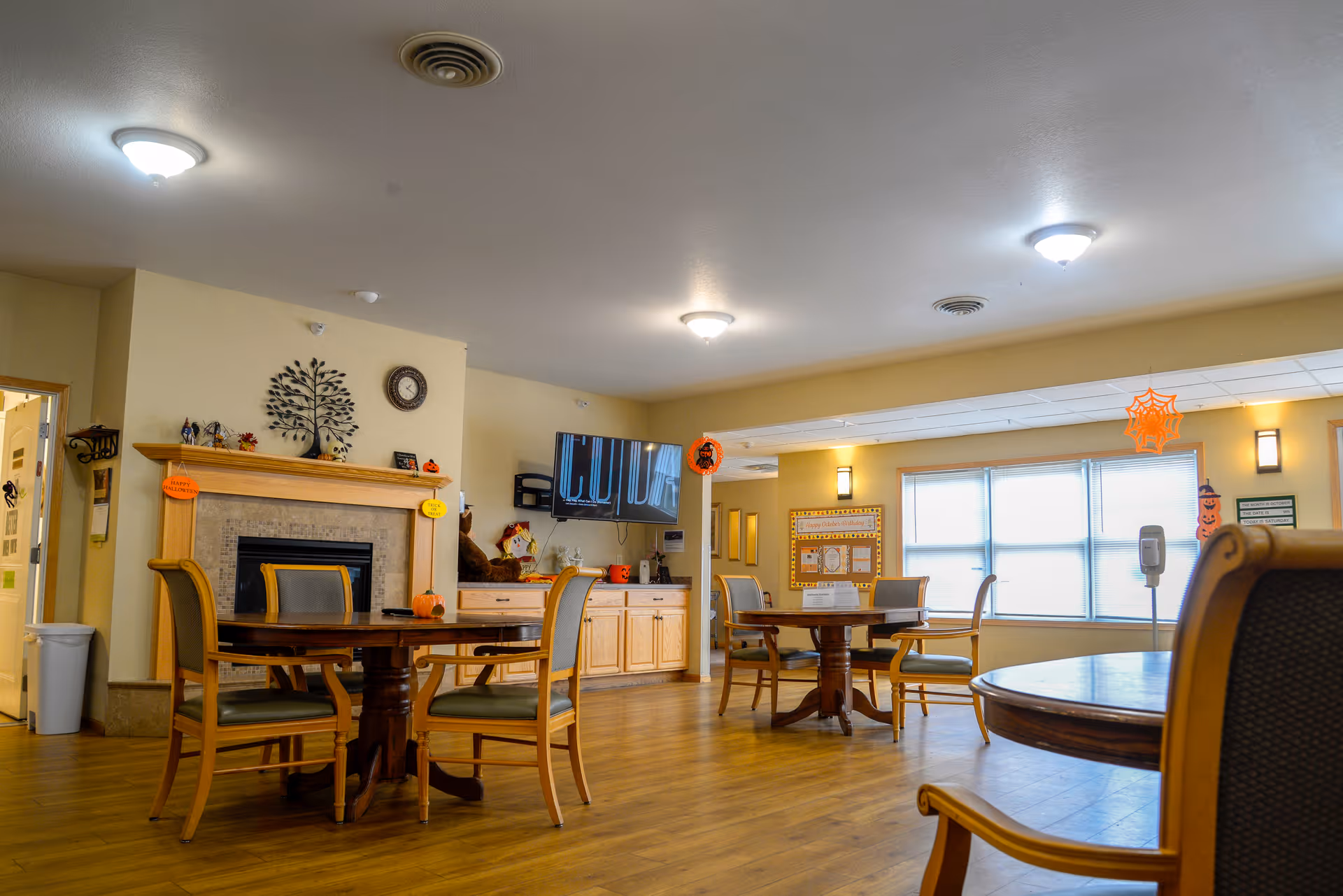 A cozy common area in a senior living facility with wooden floors, round tables, and chairs. The room features a fireplace with decorative items on the mantle, a wall-mounted TV, and large windows letting in natural light. Halloween decorations are hanging from the ceiling and walls, adding a festive touch.