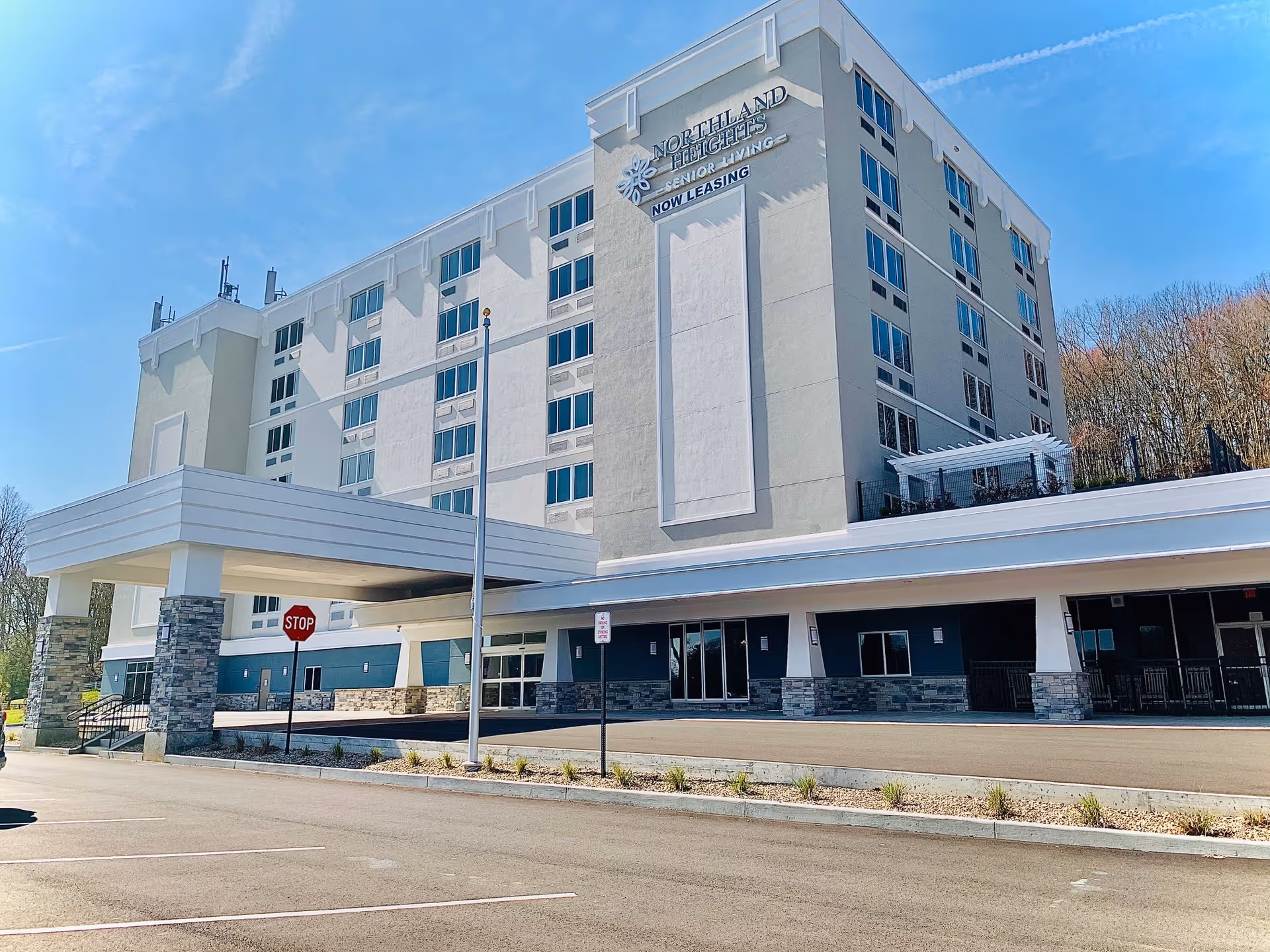Exterior view of a multi-story senior living facility building with a covered entrance and parking area in front. The building has multiple windows and a sign that reads 'Northland Heights Senior Living NOW LEASING'. Trees and a clear blue sky are visible in the background.