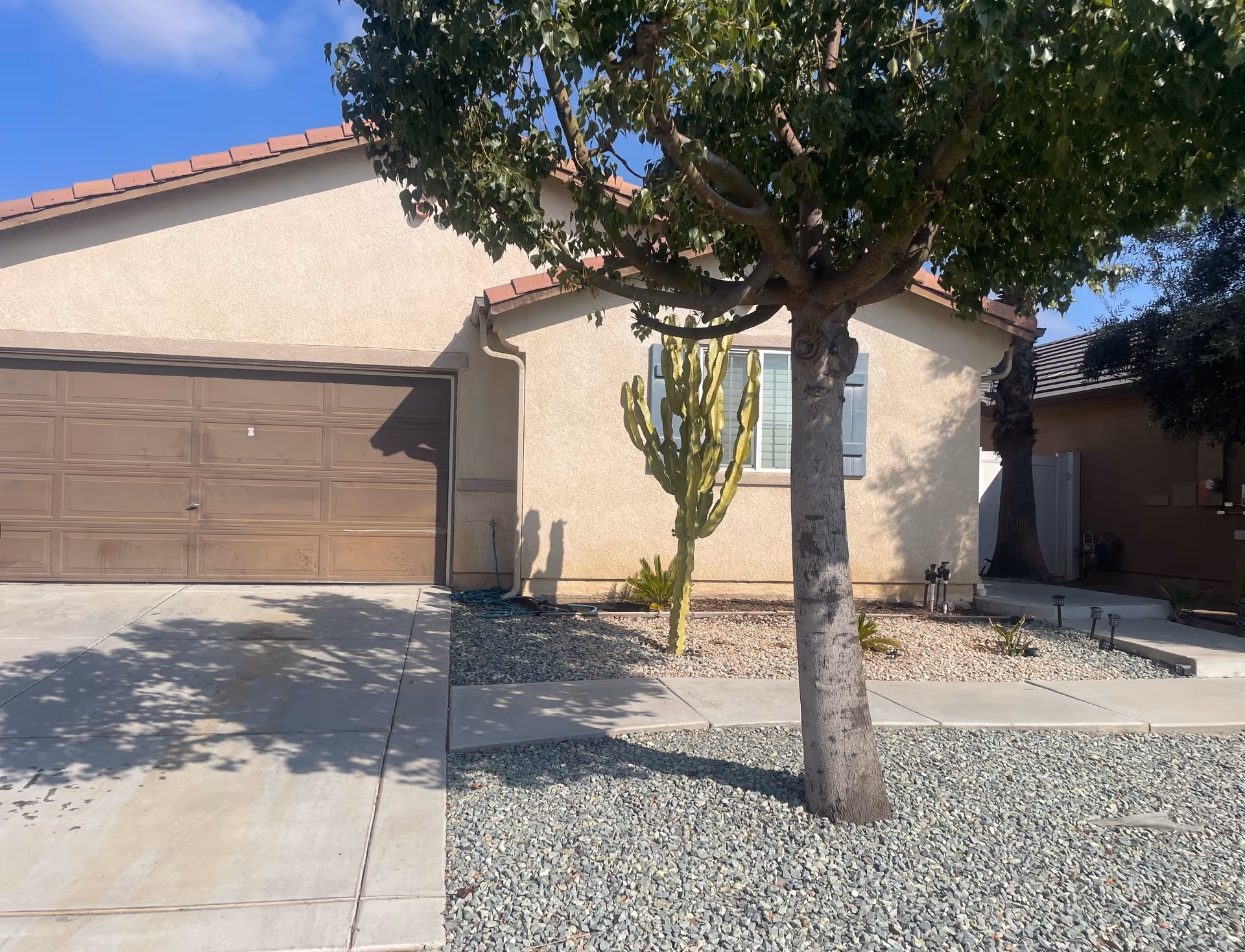 Front exterior of a single-story house with a two-car garage, a tree and a cactus in a gravel yard.