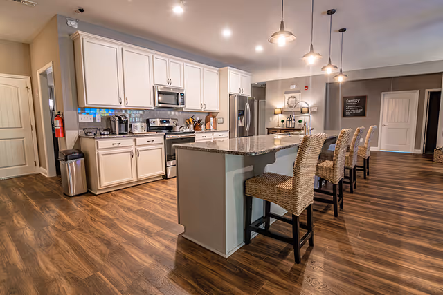 Open modern kitchen with a long granite island, four wicker barstools, white cabinets, stainless steel appliances, and wood floors.