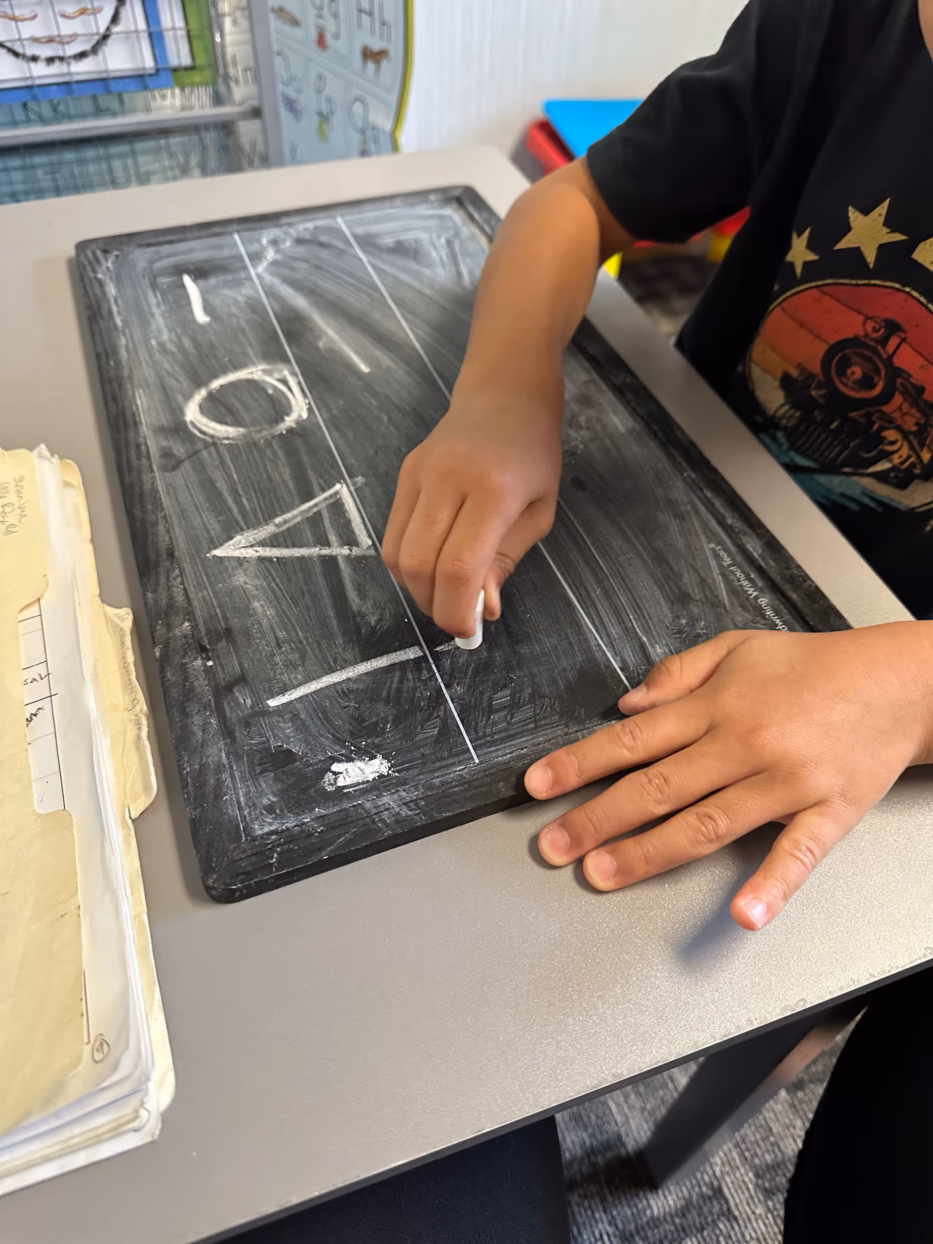 Child's hands draw on a small chalkboard at a desk indoors.