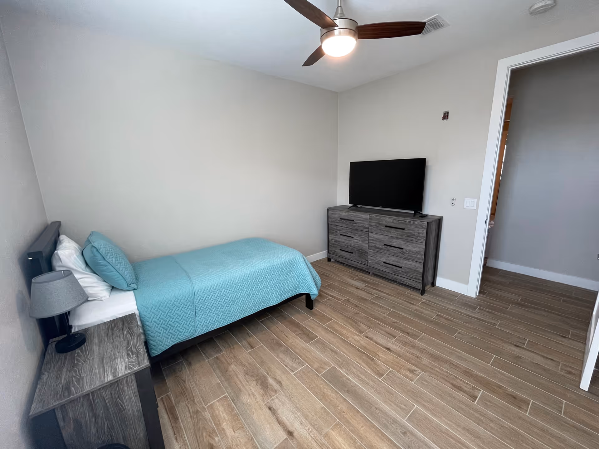A furnished bedroom with bed, dresser, nightstand, TV, and ceiling fan.