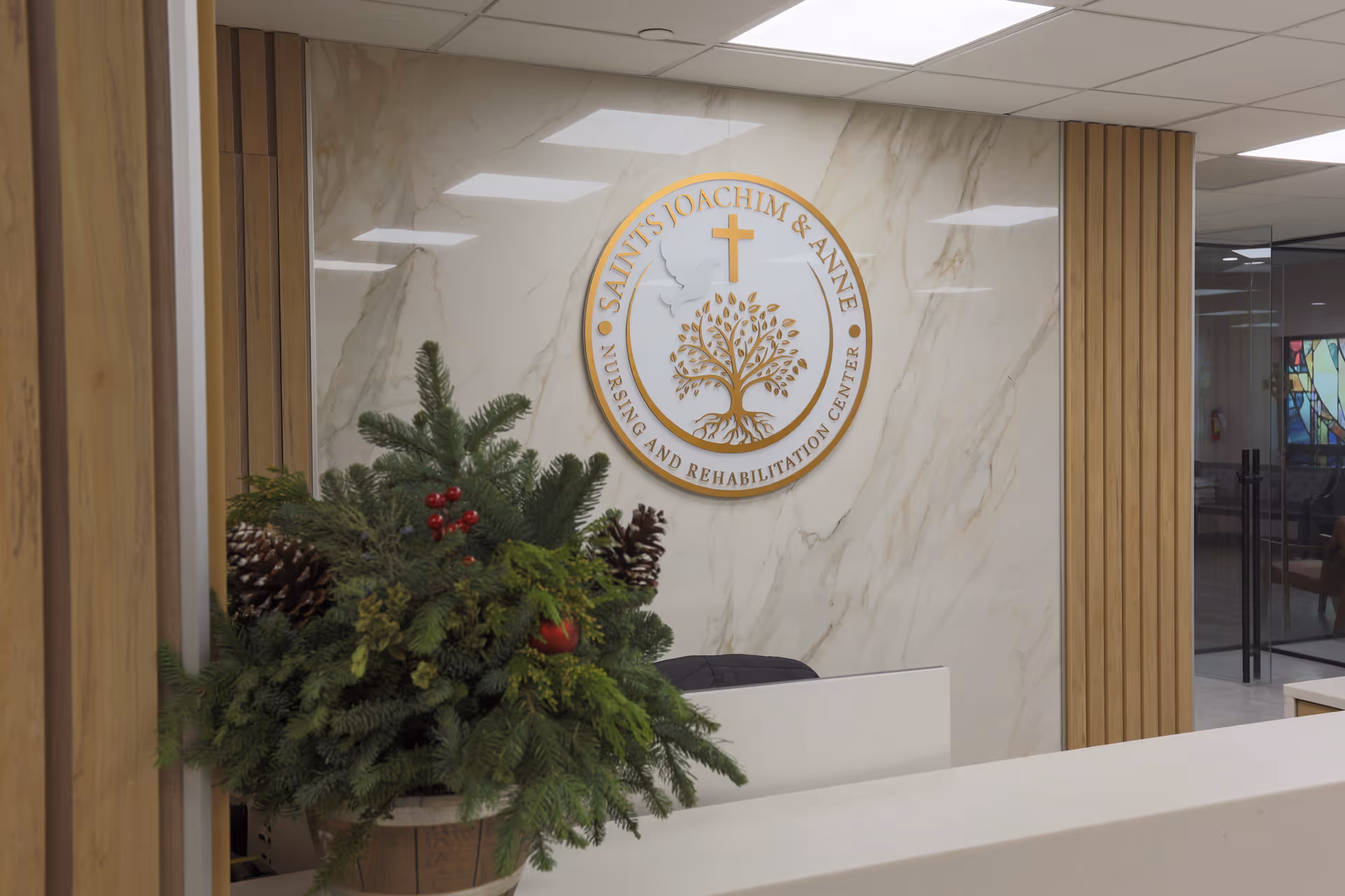 Reception area of Saints Joachim & Anne Nursing and Rehabilitation Center with a white marble wall featuring the center's logo, a wooden panel accent, and a festive green plant arrangement with pine cones and red berries on the counter.