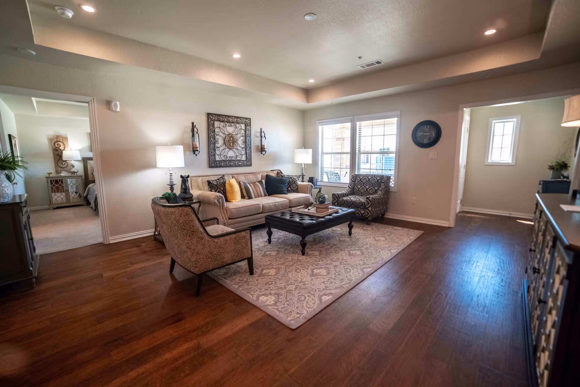 A cozy living room with a beige sofa adorned with multiple cushions, two patterned armchairs, a dark wooden coffee table on a large area rug, and two table lamps on side tables. The room has wooden flooring, a large window letting in natural light, and decorative wall art above the sofa. Adjacent rooms are visible, including a bedroom and a hallway.