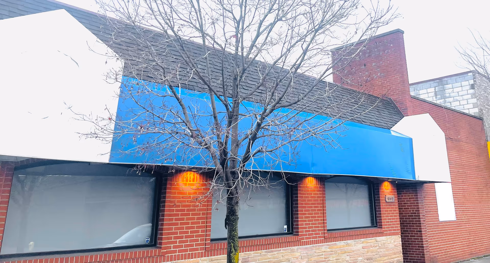 Brick building exterior with a blue awning and a leafless tree in front.