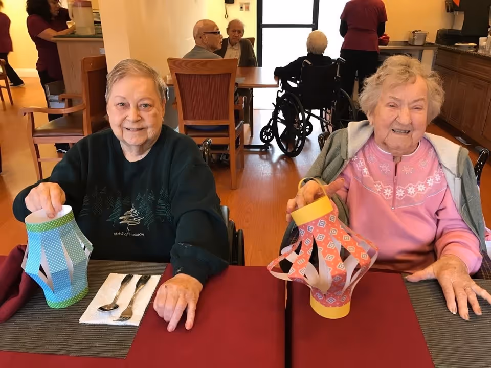 Two elderly women sitting at a dining table in a senior living facility, each holding a colorful paper lantern. There are utensils on napkins in front of them, and other residents and staff are visible in the background.