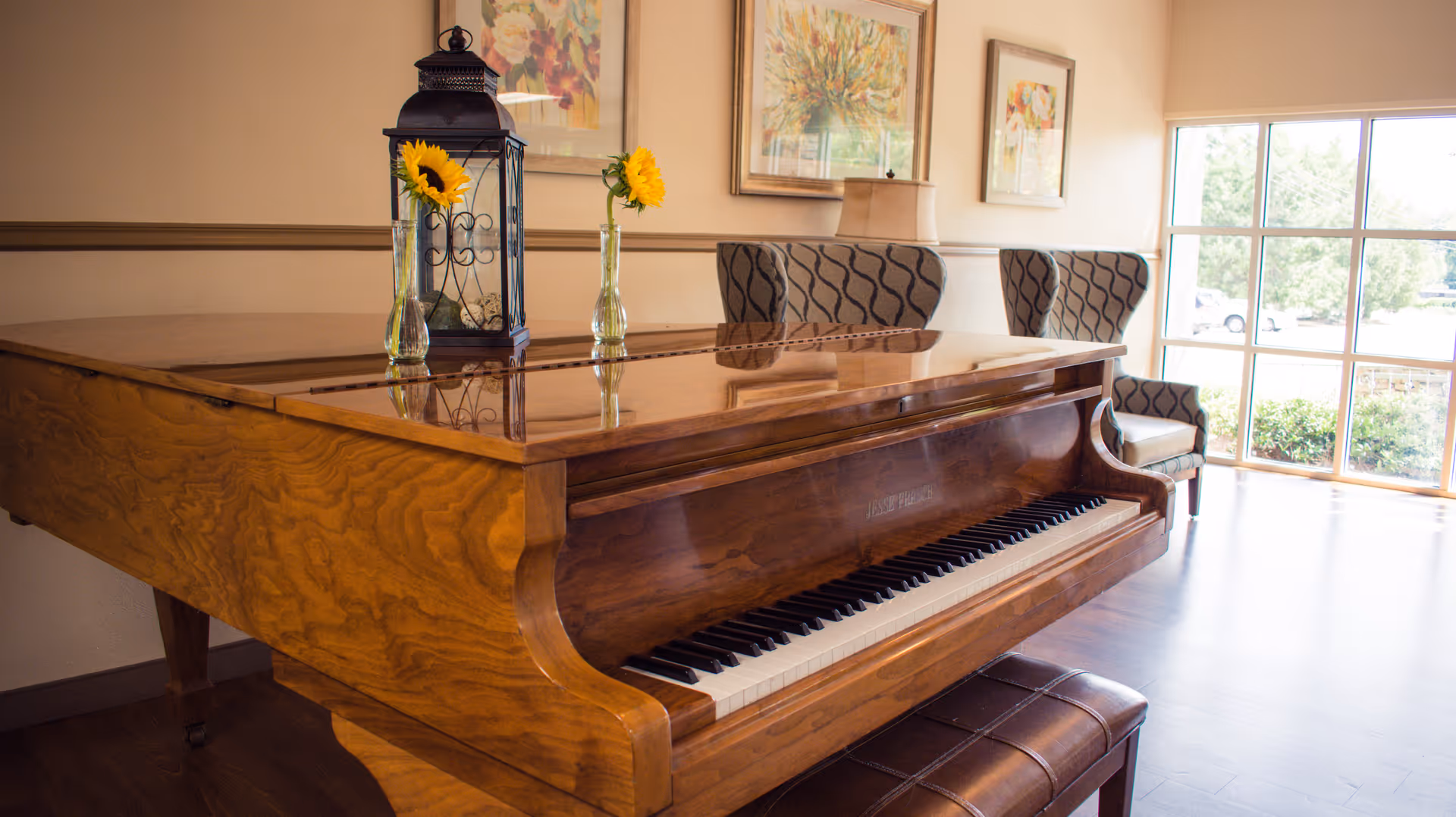 A polished wooden grand piano in a sunlit lounge with upholstered chairs, framed artwork, and vases holding sunflowers.