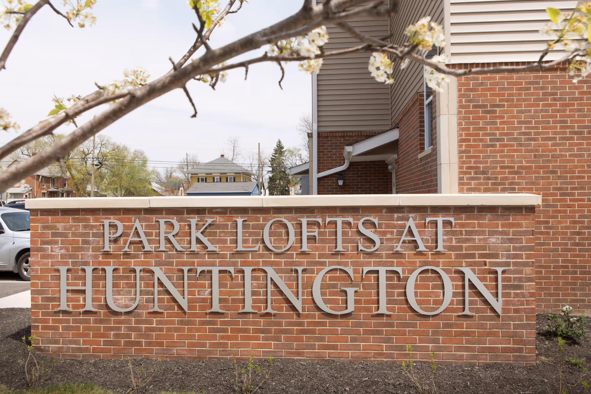 A brick sign with large silver letters that reads 'PARK LOFTS AT HUNTINGTON' in front of a building with brick and siding exterior. There are some tree branches with white blossoms in the foreground.