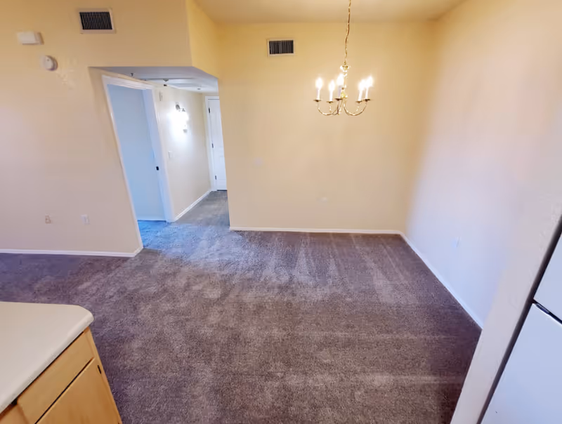 An empty interior space with beige walls, carpet, chandelier, and hallway to kitchen.